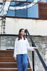 Pensive teen girl in white jacket and jeans goes down stairs in industrial urban district, thought looking away. Teenager hipster lady 13 year old outdoors. Youth gen z concept. Copy ad text space