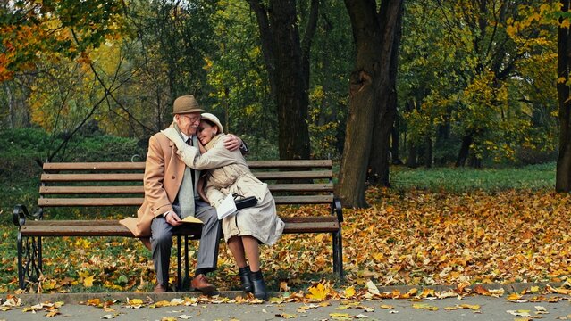 Elderly couple sitting reading a letter together on a bench in an autumn park then smiling and hugging each other