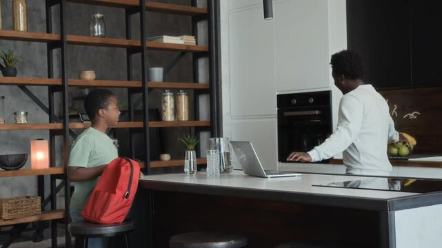 African American man see off his son to school and throws him apple in kitchen in the morning. Black skinned boy say goodbye and runs away, father works using laptop, as entrepreneur