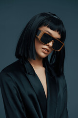 Blackhaired woman in black suit and blazer wearing sunglasses, posing against a dark blue background