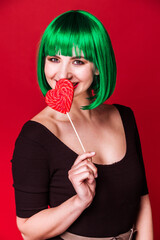 Woman with Green Hair Smiling Behind Heart-Shaped Lollipop. Young Caucasian woman with short green hair and a black top, playfully smiling behind a red heart-shaped lollipop against a red background. 