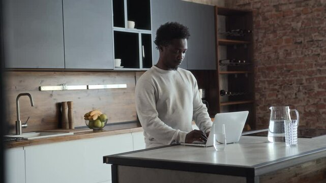 African American man is standing at kitchen table, working remotely alone using laptop in the morning. Male black skinned freelancer is typing on computer keyboard at home