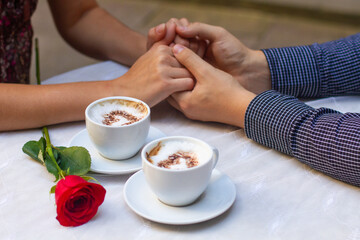 a couple in love holding hands, two cups of coffee with hearts drawn on foam and a red rose