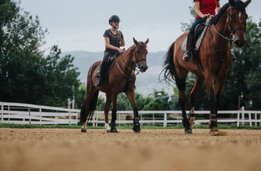 Obraz premium Two girls riding horses in an outdoor arena, showcasing equestrian skills and the bond between rider and horse on a cloudy day.