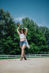 Young woman in a hat and fringe top enjoying a sunny day outdoors, standing confidently and smiling with a clear blue sky and greenery in the background.