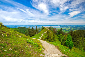 Ein Wanderweg verläuft entlang eines Bergrückens zum Chiemsee mit malerischer Sicht auf die darunter liegende Landschaft und den klaren, bewölkten Himmel, Chiemgau, Bayern, Deutschland