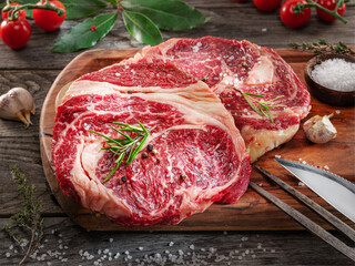 Raw beef steaks with herbs and spices ready for cooking are on wooden board.
