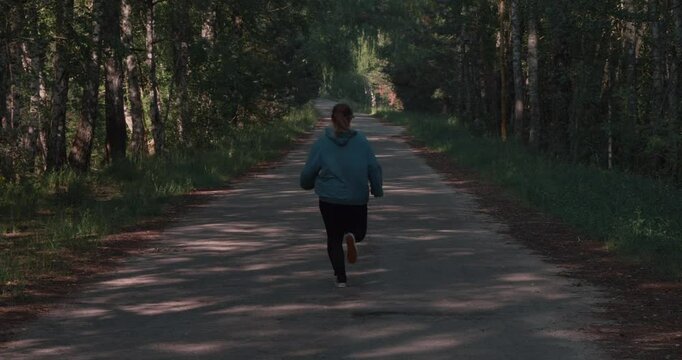 An overweight woman runs along an asphalt road through the forest. Motivation. A woman goes in for sports to lose weight and get her body in shape. Overweight woman jogging.