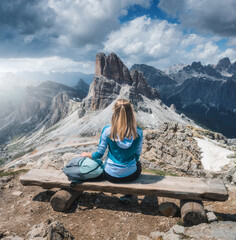 Girl with backpack is sitting on the wooden bench on mountain peak in beautiful alpine mountains at sunset in summer. Landscape with young woman in alps, high rocks, sky with clouds. Dolomites, Italy