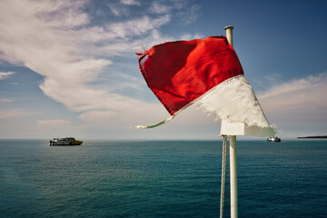 A old tattered Indonesian flag waves in the wind against a serene blue ocean backdrop. Two boats float on the horizon under a partly cloudy sky, showcasing a tranquil maritime scene.