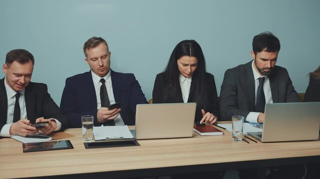 Group of business professionals seated at table during break, each engrossed in their smartphones. Prevalence of mobile addiction and distraction in corporate setting. Concept of work break