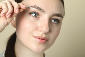 Young woman plucking eyebrow with tweezers on beige background, closeup
