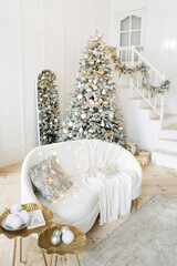Modern living room with a staircase, a Christmas tree decorated in silver and gold, white sofa with cushions and a blanket, gifts under the tree. The room exudes holiday warmth and cheer