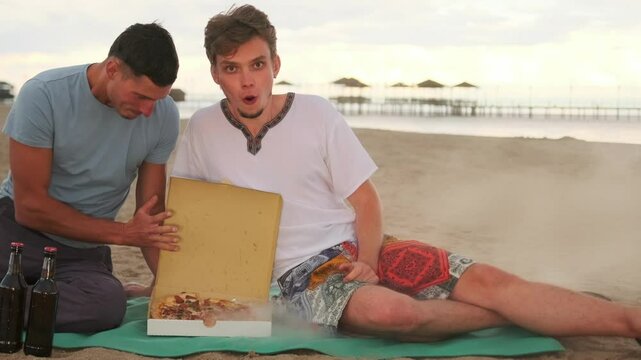 Two friends admiringly opening a box with hot pizza lying on a picnic mat on the sand near the sea.
