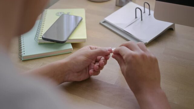 Over-shoulder shot of hands of unrecognizable young deaf male student assembling parts of prosthetic cochlear ear implant after regular cleaning, on desk with college notebooks and smartphone