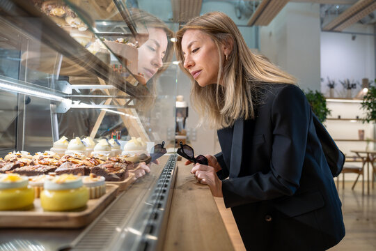Smiling business woman chooses cream and chocolate pastries at cafe display, indulging in sugary treats, looking at sweets. Sugar-free desserts and variety of cupcakes in bright confectionary window - Powered by Adobe