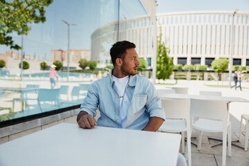 Fototapeta premium Young man sitting alone at an outdoor cafe table, looking away thoughtfully, reflecting on his thoughts on a sunny day
