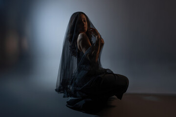 A mysterious woman in black, folds of a veil hide her face, portrait on a dark background