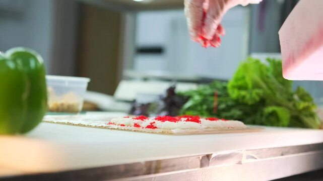 While preparing sushi rolls close-up, the chef's hand sprinkles Masago red caviar over rolled rice on bamboo mat.