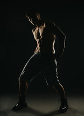 Fototapeta premium Muscular young man posing shirtless in dark studio with dramatic side lighting, emphasizing his physique and strength.