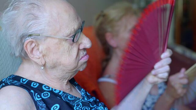 A 90-year-old woman fans herself on a hot day, with another mature woman in the background doing the same. The scene highlights the intense heat and their efforts to stay cool.