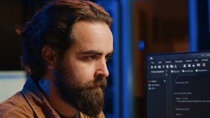 Software technician in apartment office deeply immersed in coding on computer at desk, close up. IT freelancer absorbed by work, focused on screen, writing lines of code at workstation, camera A
