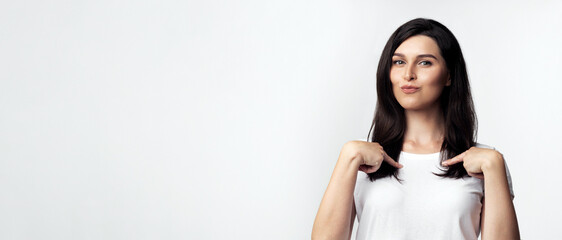A young woman wearing a white t-shirt is pointing to herself with both hands in front of a white...