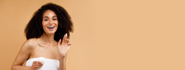 Naklejka premium Skin care concept. Pretty african american lady applying moisturizing face cream, posing wrapped in towel and smiling at camera, standing over brown background, copy space