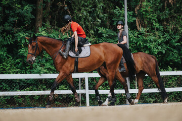 Two young girls ride horses in an outdoor arena, enjoying an equestrian activity surrounded by green foliage