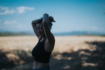A young woman enjoying a sunny day outdoors in a scenic field, tying her hair up. The atmosphere is relaxed and carefree.