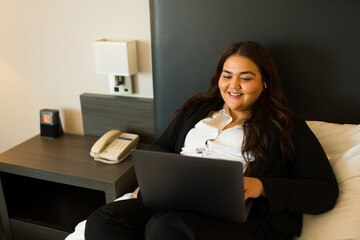 Hispanic woman in business attire sits on a hotel bed, using a laptop with earphones