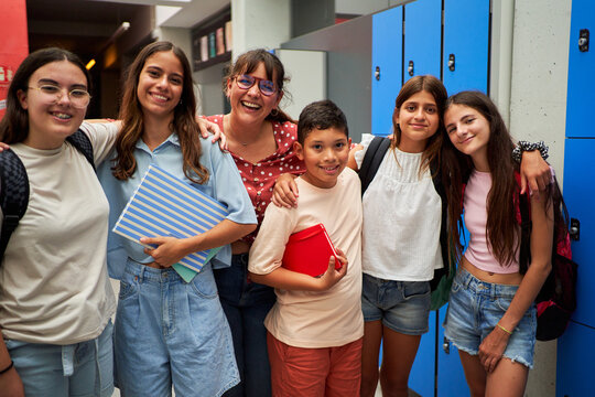Cheerful portrait of a group of preteen student friends with their female teacher inside the school looking at camera smiling and embracing to each other.