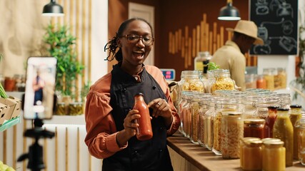 African american vendor promoting homemade sauces and honey, creating online video advertisement to present eco friendly products at zero waste supermarket. Woman recording promotional ad. Camera 1.