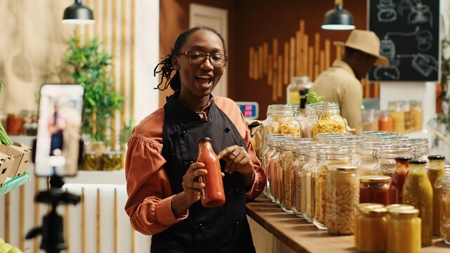 African american vendor promoting homemade sauces and honey, creating online video advertisement to present eco friendly products at zero waste supermarket. Woman recording promotional ad. Camera 1.