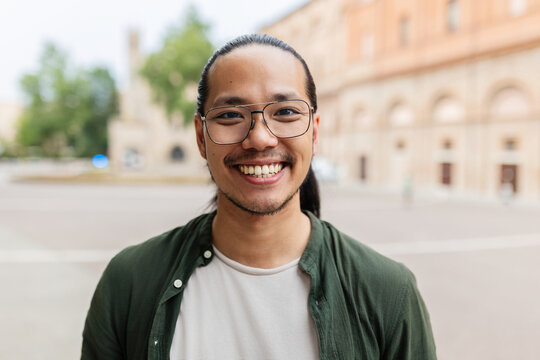 Smiling portrait of young asian man looking at camera standing at city street. Joyful millennial chinese male feeling positive, closeup outdoor headshot. Happy people