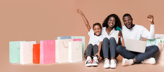 Online shopping for family. Happy african american parents and daughter sitting on floor with laptop, credit card and bright shopper bags.