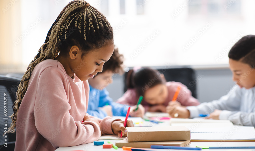Poster portrait of small black girl with afro braids sitting at table in classroom at primary school or kin