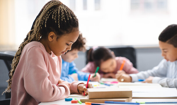 Portrait of small black girl with afro braids sitting at table in classroom at primary school or kindergarten, writing or drawing in notebook. Reopening and return back to school after lockdown - Powered by Adobe