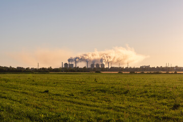 Fototapeta premium A grass field, on the horizon there is a working chemical plant, there are many clouds of smoke from the factory chimneys, illuminated by sunlight. Ecological concept image in summer during sunset