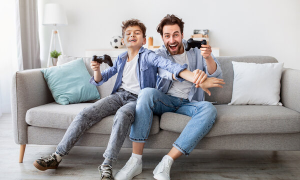 Joyful dad and son with joysticks playing video games at home, boy distracting father with hand, faving fun together, sitting on sofa in living room. Fatherhood and leisure activities concept