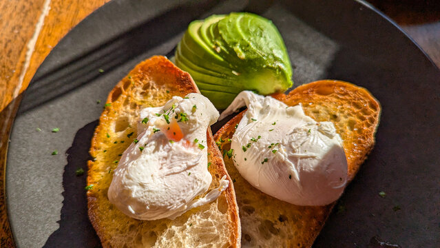 Close up of two poached eggs on sourdough toast with a side of avocado on black grey plate for breakfast brunch in beautiful sunlight at local cafe