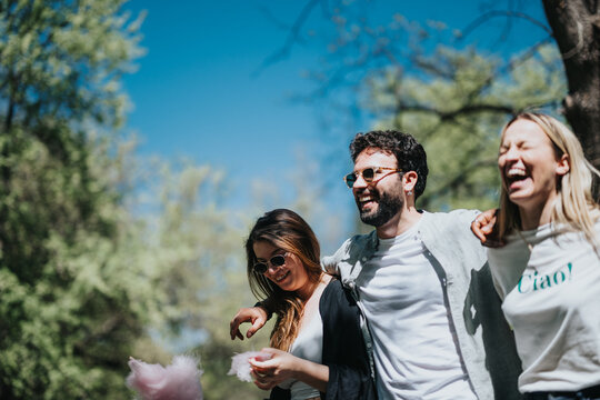 Three diverse friends share a moment of joy and laughter at the park while enjoying cotton candy on a sunny spring day, promoting a sense of casual fun and togetherness.
