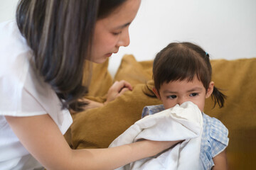 Woman helps young girl blow her nose with tissue on a couch. Child is comforted by the woman during illness or cold.