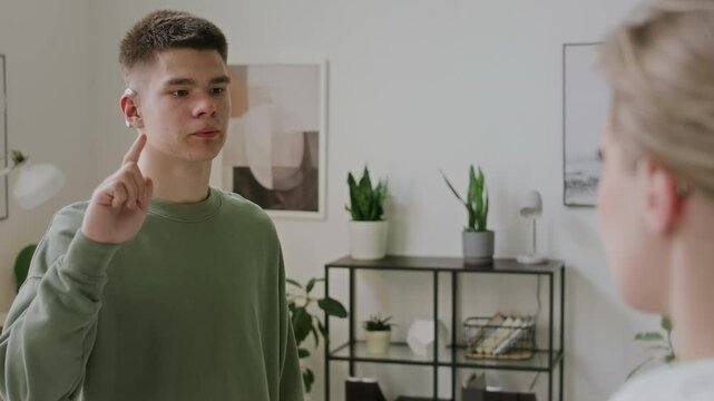 Medium over-shoulder shot of Caucasian male teenager with cochlear implant standing in classroom with female teacher, reciting sign language alphabet letters in ASL, and teacher nodding