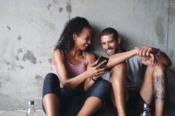 Man, woman and fitness break with phone for exercise podcast, streaming in building for wellness. Couple, mobile app or workout together with rest in parking lot for health, video for gym or training