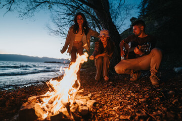 Fototapeta premium Group of three friends gathered around a warm campfire on the beach, enjoying live guitar music under an evening sky.