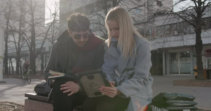 Seated outdoors under sunlight, business colleagues review documents on a digital tablet, discussing tasks in a casual setting, blending work with a refreshing environment.