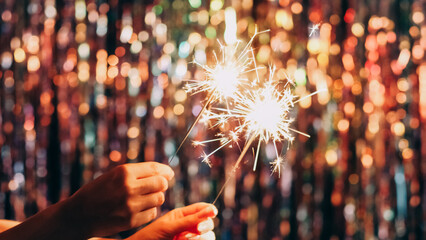 Christmas sparklers. Winter holidays. Festive mood. New year party. Female hands holding burning bengal fireworks on defocused glow bokeh lights tinsel decor background.