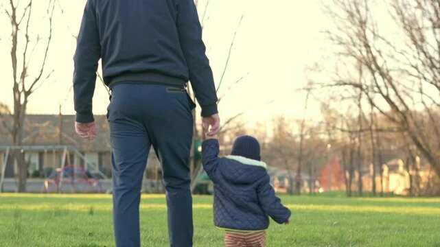 A father and his toddler walk hand in hand through a grassy park on a sunny day. The video captures the warmth and bonding of a family outing, highlighting the joy of spending time together outdoors.