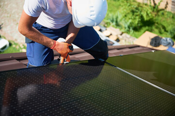 Worker building photovoltaic solar panel system on rooftop of house. Close up of man engineer in helmets and gloves installing solar module with help of hex key outdoors. Alternative, renewable energy